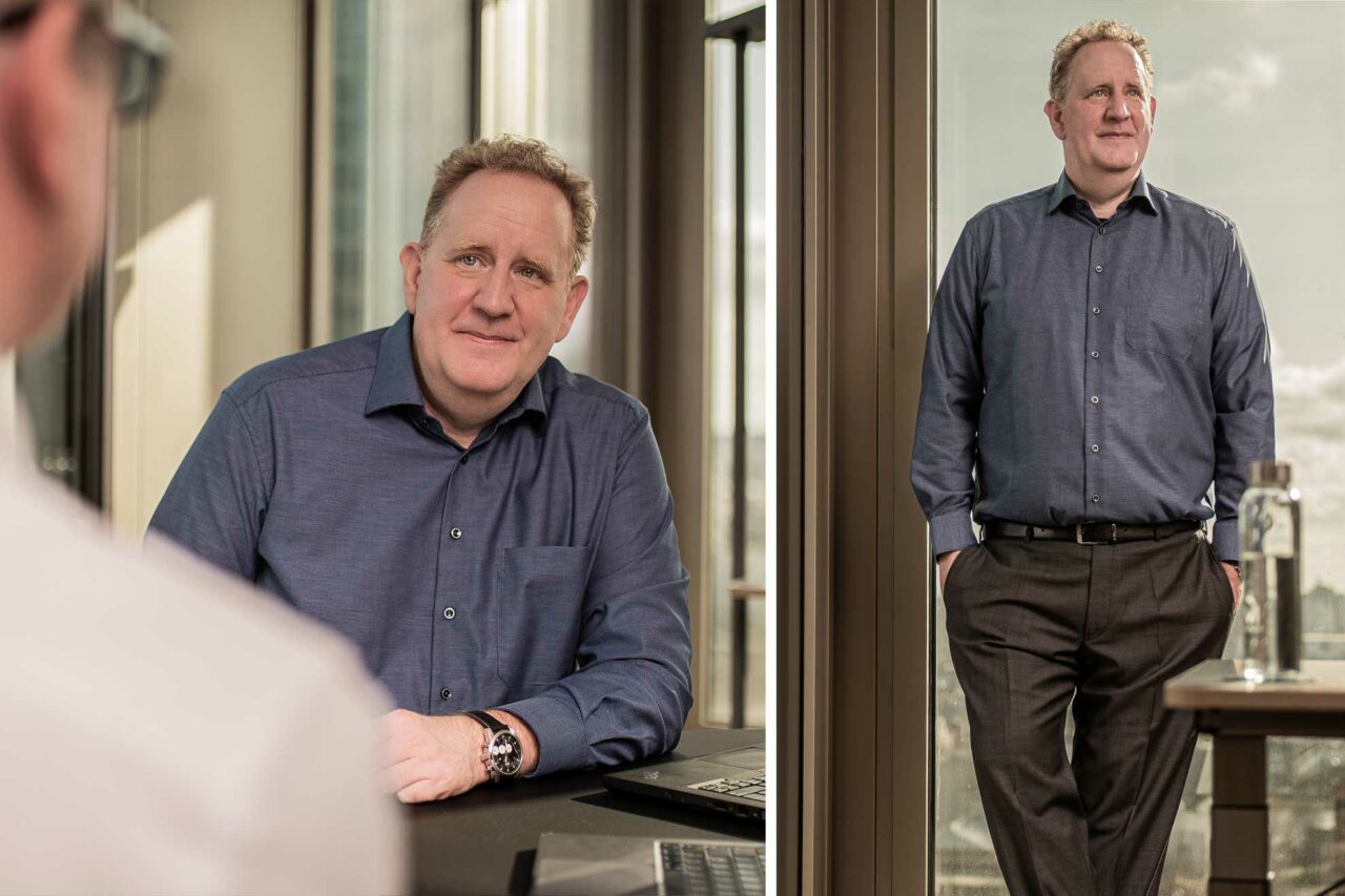 Businessfotos für Franke Private Finance. Ein professionelles Business-Porträt eines Mannes in einem blauen Hemd, aufgenommen in einem modernen Büro. Auf der linken Seite sitzt er an einem Schreibtisch und blickt freundlich in die Kamera, während im Hintergrund eine Büroumgebung zu sehen ist. Auf der rechten Seite steht er entspannt an einem Fenster mit verschränkten Händen in den Hosentaschen, mit einem Lächeln auf dem Gesicht. Das natürliche Licht fällt sanft durch das Fenster und verleiht der Aufnahme eine einladende, professionelle Atmosphäre.