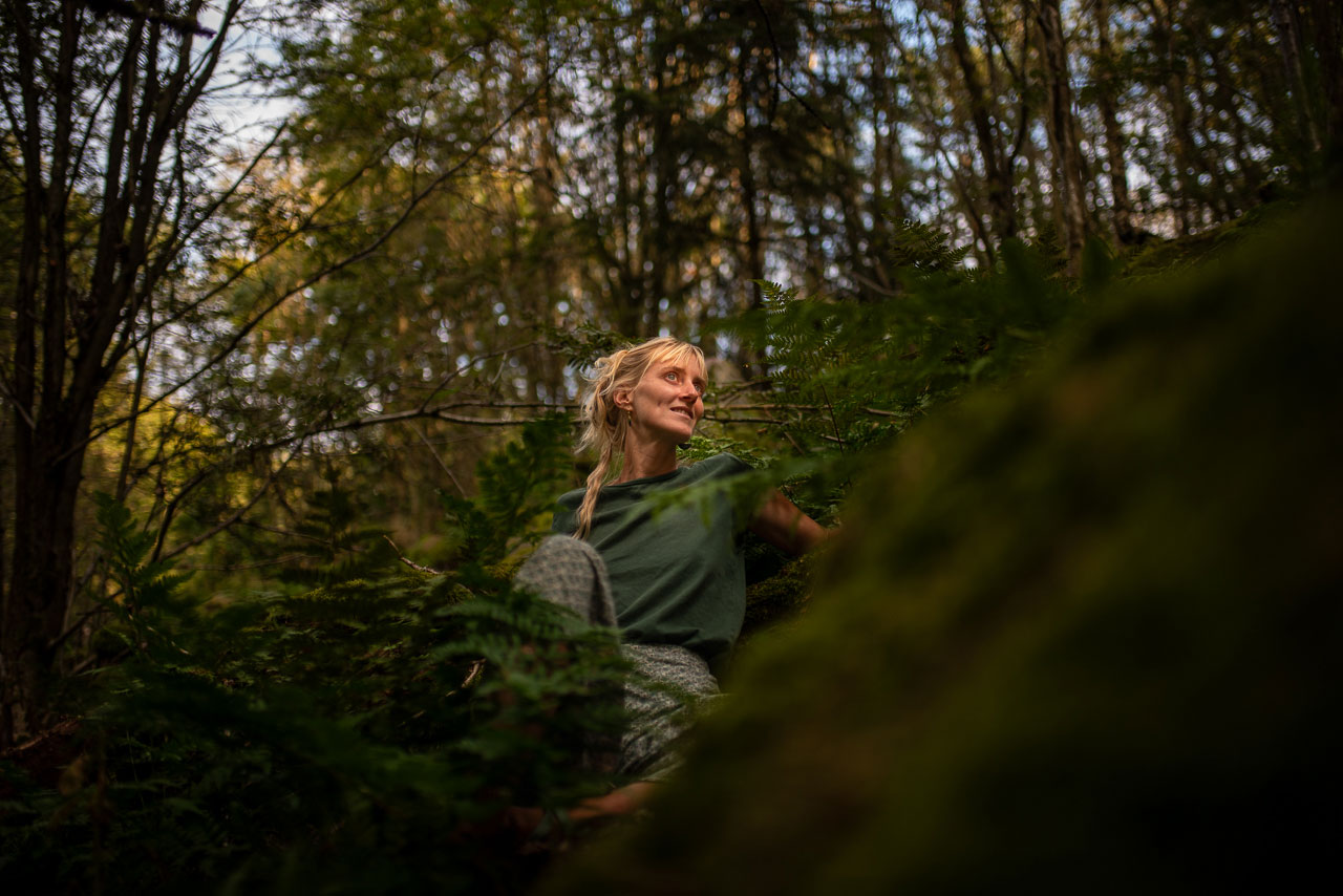 Eine junge Frau in grüner Kleidung sitzt entspannt im Wald, umgeben von Farnen und Moos. Sie blickt lächelnd zur Seite, während das warme Licht die natürliche Umgebung hervorhebt und eine ruhige, harmonische Atmosphäre schafft.