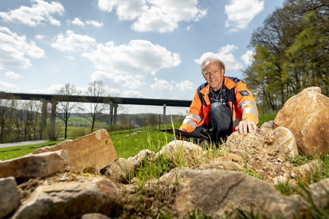 Mitarbeiter in orange-blauer Arbeitsjacke kniet neben Felsen im Freien, mit einer Brücke im Hintergrund. Er arbeitet an einem Außeneinsatz bei sonnigem Wetter.