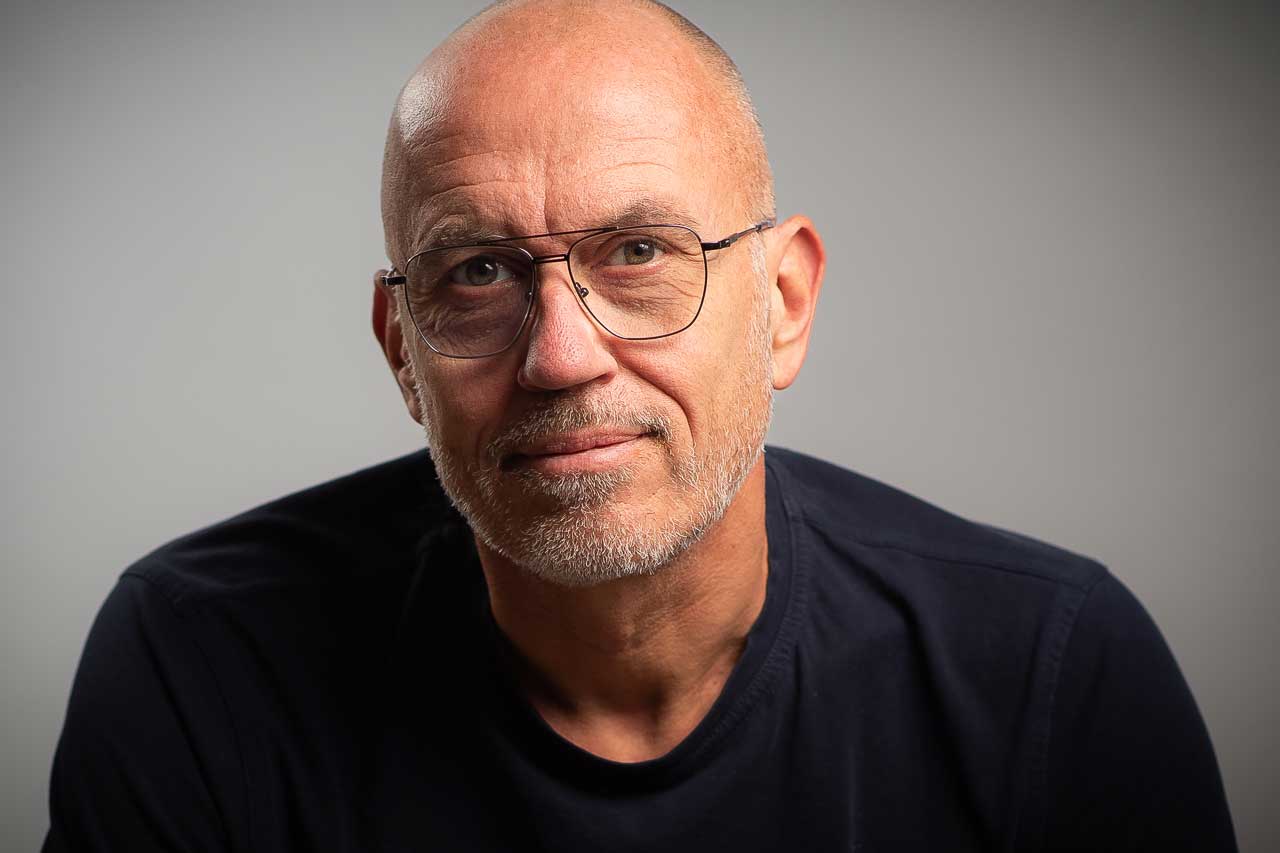 Authentisches Porträt eines älteren Mannes mit Brille, direkter Blick in die Kamera, ruhiger Hintergrund – symbolisiert Selbstbild und Haltung.