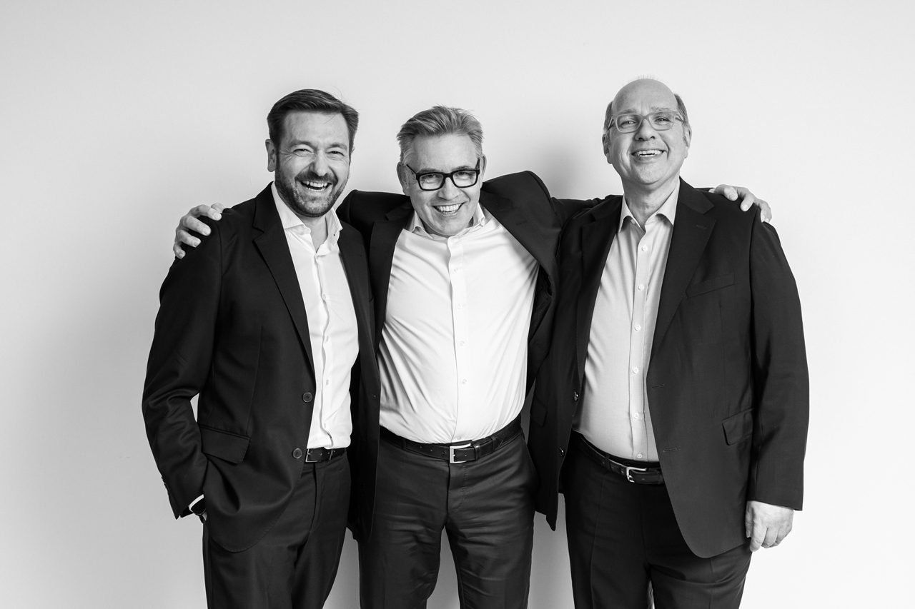 Drei Männer im Anzug lachen, stehen eng zusammen und legen die Arme umeinander vor hellem Hintergrund.